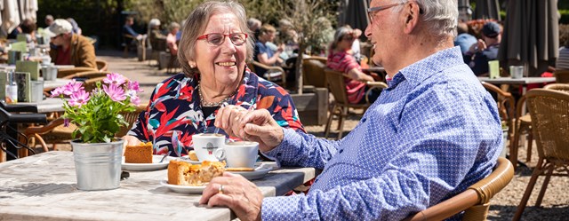 Ouder echtpaar zit op een zonnig terras met koffie en appeltaart.