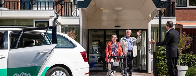 Echtpaar loopt naar buiten bij de woning en zwaait naar de Avan chauffeur hen op komt halen met een personenauto.